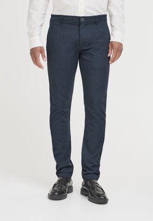 Homme portant un pantalon slim bleu marine foncé, une chemise blanche à manches longues et des mocassins en cuir noirs, debout sur un fond blanc.