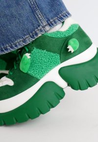 Sneakers chunky verdi e bianchi con materiali testurizzati e dettagli giocosi, parzialmente coperti da jeans in denim, su uno sfondo chiaro.