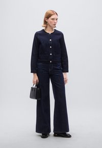 Ναυτικό τζιν μπουφάν με στρογγυλή λαιμόκοψη και πέντε ασημένια κουμπιά, σε συνδυασμό με φαρδιά μπλε τζιν παντελόνια. Το μοντέλο κρατά μια μικρή μαύρη τσάντα χειρός.