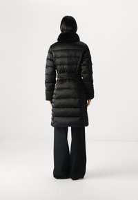 Zwarte pufferjas met een getailleerde taille en nep bontkraag. Kenmerkt zich door een gestikt ontwerp en een iets uitlopende silhouet, dat tot halverwege de kuit reikt.