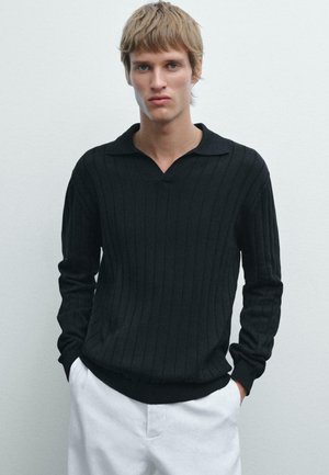 Schwarzer, gerippter Strickpullover mit V-Ausschnitt und langen Ärmeln, kombiniert mit weißen Hosen. Glatte Textur, lässige Passform.