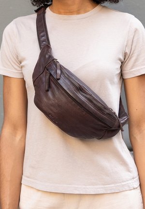 Person iført en lysebeige t-shirt med en mørkebrun læder crossbody-taske med lynlås hen over brystet mod en grå baggrund.