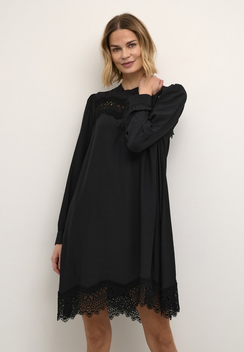 Schwarzes Kleid mit langen Ärmeln, rundem Ausschnitt und filigranen Spitzenverzierungen am Saum, das eine fließende Silhouette und weiche Textur zeigt.
