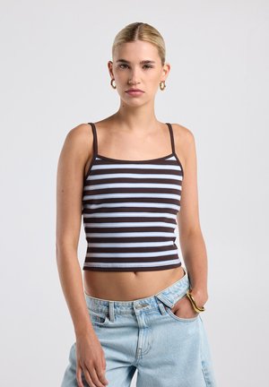 Jonge vrouw die een zwart-wit gestreept tanktopje en lichtblauwe spijkerbroek draagt, met één hand in de zak tegen een effen achtergrond.