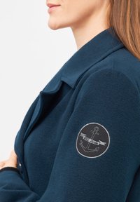 Giacca in pile blu navy con colletto pieghevole e toppa ricamata nera che presenta un'ancora e un banner sulla manica sinistra.