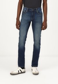 Jeans en denim bleu foncé avec une coupe slim, dotés de cinq poches et de détails de couture subtils. Associés à des baskets blanches et grises.