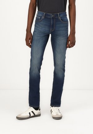 Jeans de mezclilla azul oscuro con corte ajustado, que cuentan con cinco bolsillos y detalles de costura sutiles. Combinados con zapatillas blancas y grises.
