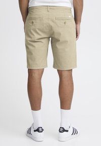 Shorts beiges avec un motif texturé, deux poches arrières avec des boutons, et une étiquette à la taille, accompagnés de chaussettes blanches et de baskets avec accents noirs.