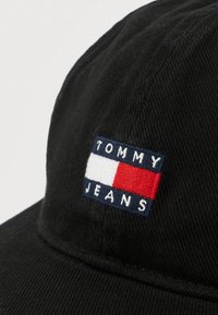 Gorra de algodón negra con un parche con el logo que presenta franjas horizontales en azul, blanco y rojo, y el texto "TOMMY JEANS" bordado en blanco.
