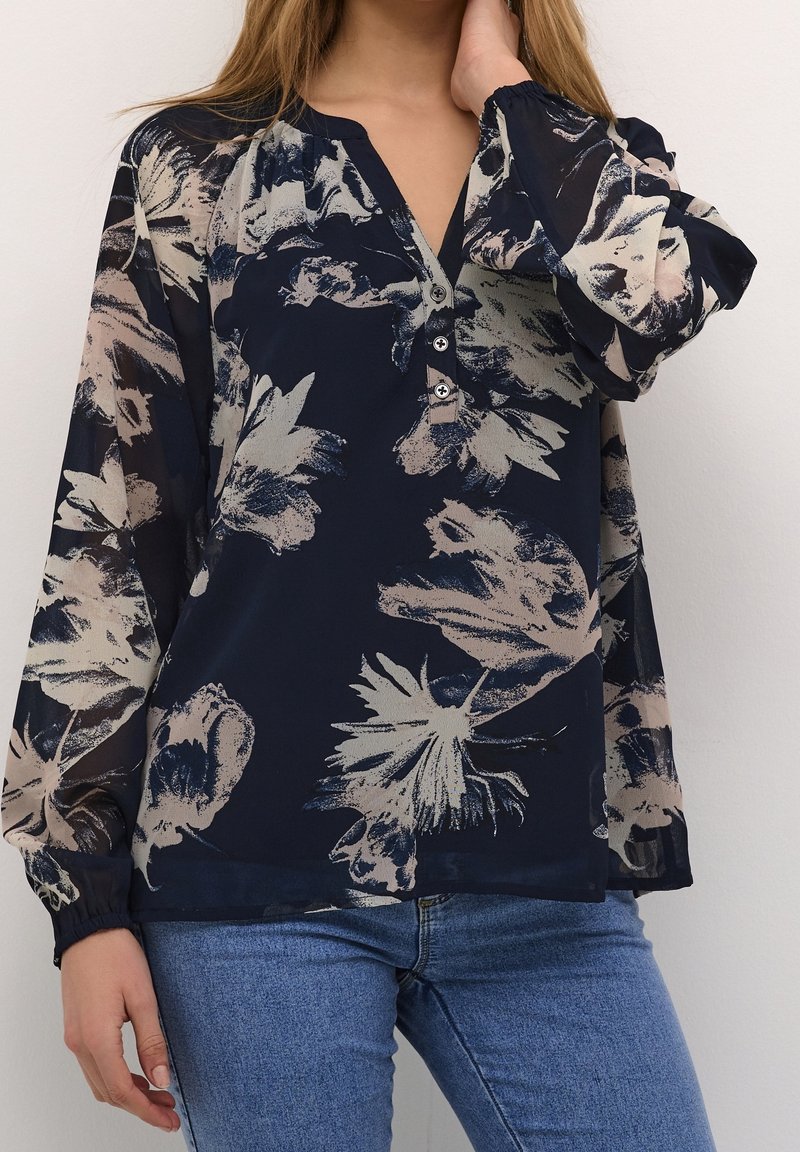 Femme portant un chemisier bleu marine à grand imprimé floral beige avec un col en V à trois boutons, associée à un jean bleu clair, main touchant le cou.