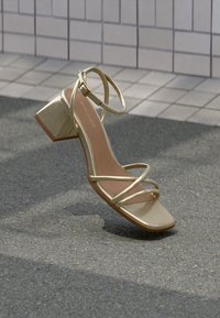 Sandale cu toc auriu metalic, cu toc pătrat, bretele subțiri și vârf rotunjit. Textură netedă cu talpă plată.
