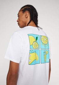 Camiseta blanca de algodón con un diseño gráfico en la parte posterior: cuatro cuadrantes con ilustraciones de limón y limonada en amarillo y azul.