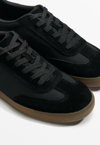 Black athletic shoes made of suede and synthetic material, featuring black laces, a smooth toe cap, and a textured gum rubber sole.