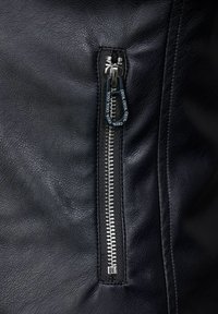 Sezione di giacca in pelle nera con zip argentata, superficie testurizzata e sovrapposizione in tessuto alla base della zip, per garantire funzionalità.