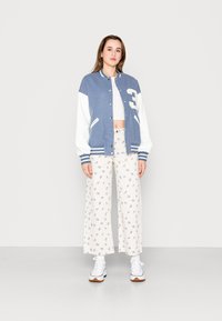 Casaco varsity azul com mangas brancas e um grande número "3", combinado com calças largas brancas com estampado floral e ténis brancos.