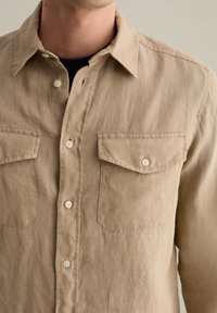 Beige linnen overhemd met een knoopsluiting, twee borstzakken en een klassieke kraag. Getextureerde stof met een natuurlijke, zachte uitstraling.