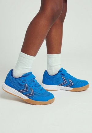 Benen met witte crew-sokken en felblauwe sportschoenen met zwart-witte chevronstrepen en rubberen zolen op een lichte achtergrond.