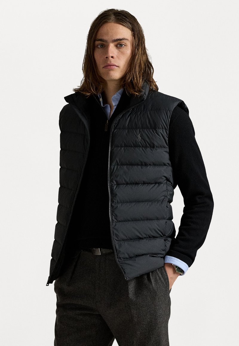 Sort puffervest med høj krave, lynlås foran og vandret quiltet design, båret over en sort sweater og en blå skjorte med krave.