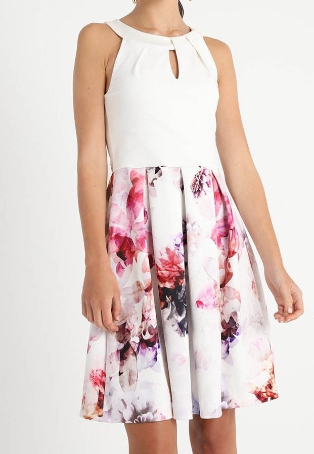 Femme portant une robe blanche sans manches avec encolure goutte d'eau et jupe plissée à motif floral rose, violet et rouge.