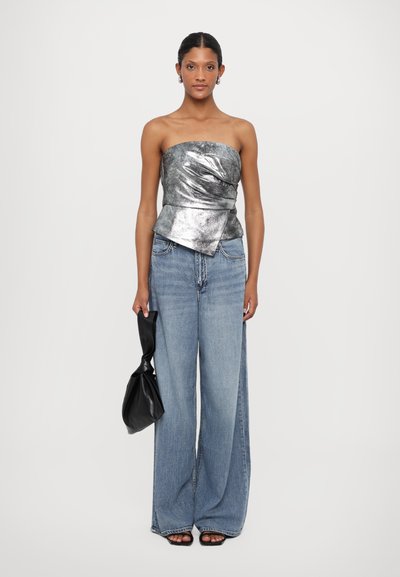 Metaliczny srebrny top bez ramiączek w stylu peplum w zestawie z szerokimi, jasnoniebieskimi dżinsami. Modelka trzyma czarną torbę z węzłowym akcentem.