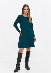 Turkusowa sukienka z długim rękawem, okrągłym dekoltem, bocznymi kieszeniami i złotymi guzikami na ramionach. Noszona z czarnymi kozakami do kolan.