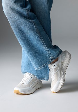 Weiße Sneaker mit Mesh-Gewebe und Gummisohle, die ein strukturiertes Muster und dezente Akzente aufweisen, kombiniert mit weit geschnittenen blauen Jeans.