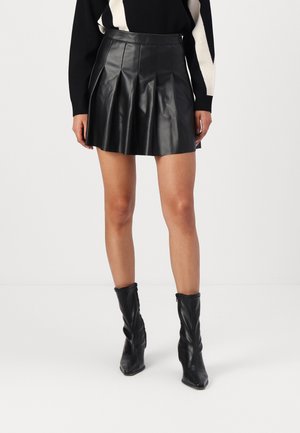 Mujer con falda mini plisada de cuero negra, suéter blanco y negro, y botas negras de tacón hasta la mitad de la pantorrilla, posando frente a un fondo liso.