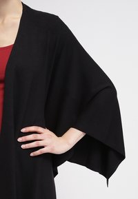 Vrouw in een zwarte sjaal over een rode top, met haar rechterhand op haar heup rustend tegen een effen grijze achtergrond.