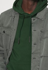 Lichtgrijs jack over een donkergroene hoodie, met een voorzak, verstelbare trekkoorden en drukknopen als sluiting.