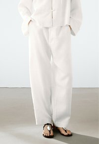 White linen wide-leg pants feature side pockets, a loose fit, and a textured surface. Paired with black sandals having silver accents.