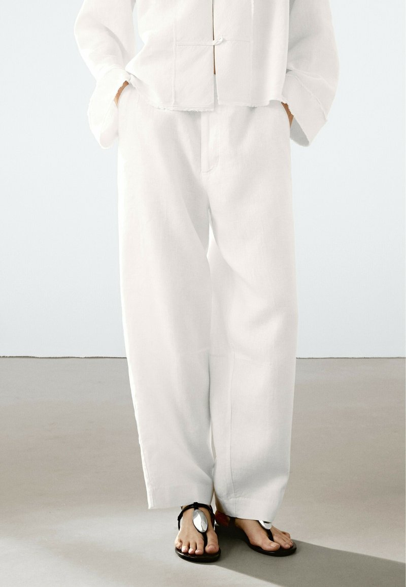White linen wide-leg pants feature side pockets, a loose fit, and a textured surface. Paired with black sandals having silver accents.