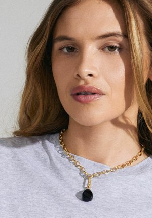 Collier en chaîne épaisse en ton doré avec un pendentif en forme de goutte noire, porté sur une chemise grise, présentant des textures lisses et des finitions polies.
