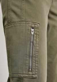 Pantalones cargo verde oliva con un bolsillo lateral con cremallera de metal plateada y costuras expuestas, confeccionados en una suave tela de algodón.