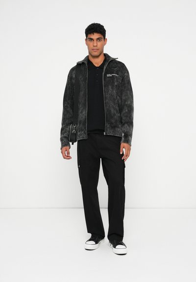 Jeune homme portant un polo noir, une veste zippée noire délavée, un pantalon cargo noir, des baskets noir et blanc, et portant un petit sac à bandoulière noir.