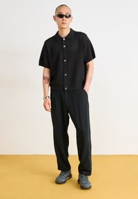 Black knit short-sleeve shirt with a collared neckline and button closure, paired with loose black trousers and gray athletic shoes.