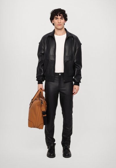 Black leather jacket and pants, white shirt, black boots. Holding a brown leather bag with adjustable strap, features minimalistic design.