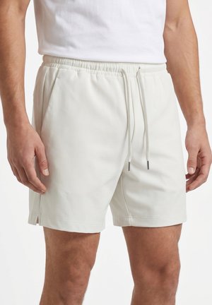 Primer plano de un hombre con pantalones cortos beige claro con cintura elástica, cordón y bolsillos laterales, combinados con una camisa blanca.