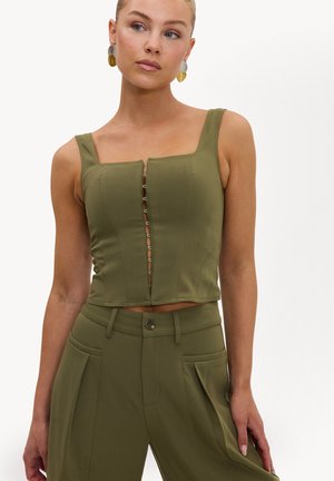 Femme portant un top court vert olive avec des crochets à l’avant et un pantalon taille haute assorti avec des poches latérales, légèrement tournée vers la droite.
