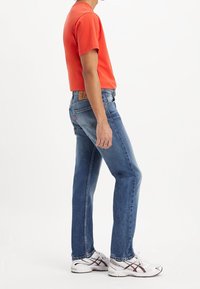 Jeans ajustados de color azul con un acabado desgastado, combinados con una camiseta de manga corta de color naranja brillante. Lleva unas zapatillas deportivas blancas.