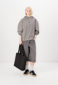 Hanorac oversized gri cu text negru, pantaloni scurți gri, geantă tote neagră, șosete albe și adidași negri cu talpă din gumă, stând pe un fundal alb.