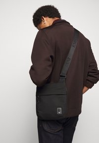 Svart axelremsväska i slät nylon med justerbar rem. Har en framflik, dragkedjestängning och en liten logopatch.