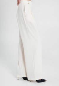 Pantaloni a gamba larga in tessuto bianco crema, con pieghe frontali, una vita sartoriale con fibbia dorata e décolleté neri a punta.