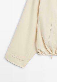 Lange crèmekleurige jas met mouwen met twee knopen aan de manchetten en een elastisch trekkoord aan de zoom, op een lichte achtergrond.