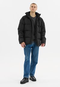 Doudoune noire avec capuche, présentant des sections matelassées et une fermeture éclair. Associée à un jean bleu et des chaussures noires à semelle épaisse.