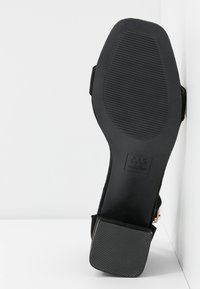 Semelle de sandale noire avec grip texturé au talon et à l'avant, inclinée verticalement contre une surface d'angle blanche.