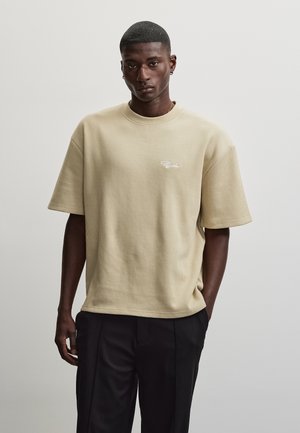 Jeune homme aux cheveux courts portant un t-shirt côtelé beige à manches courtes et un pantalon noir, debout devant un fond clair uni.