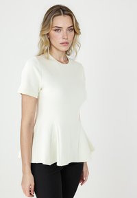 Top peplum in crema chiaro con maniche corte e scollatura rotonda. Presenta un orlo svasato e tessuto testurizzato. Abbinato a pantaloni neri.