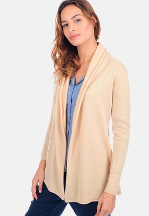 Cardigan beige avec un devant drapé ouvert, des manches longues, une texture douce et sans fermeture, superposé sur une chemise bleu clair.