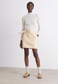 Haut à col roulé crème et jupe en cuir beige mini avec taille ceinturée. Modèle portant des chaussettes légères et des mocassins beiges sur un fond neutre.