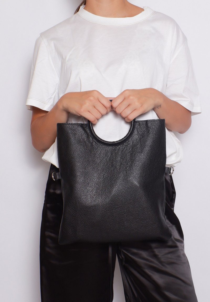 Borsa tote in pelle nera con una finitura testurizzata, dotata di un manico superiore arrotondato e un design minimalista, tenuta da una persona in camicia bianca.